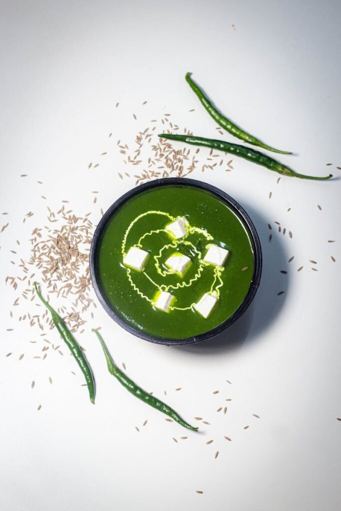 Top-down shot of a vibrant green soup with paneer cubes and cumin seeds on a white background.