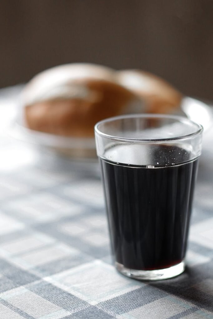 A refreshing glass of dark soda on a checkered tablecloth with bread in the background.