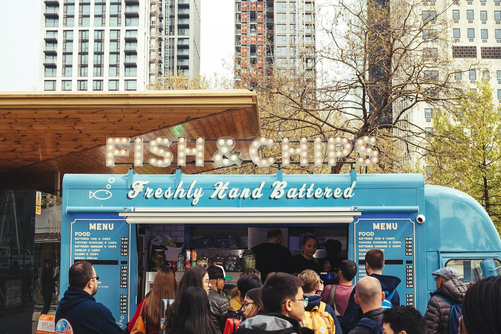 Crowd enjoying fish and chips at a lively food truck with urban buildings in the background.