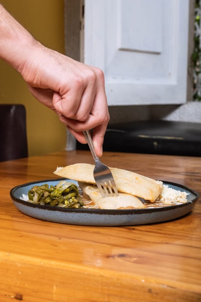 A hand holds a fork, cutting into tamales served with vegetables on a wooden table.