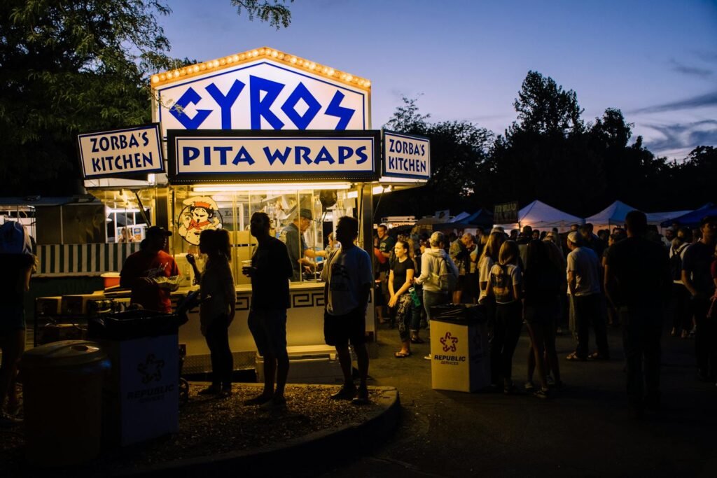 Crowds gather at a food festival stall serving gyros and pita wraps during the evening.