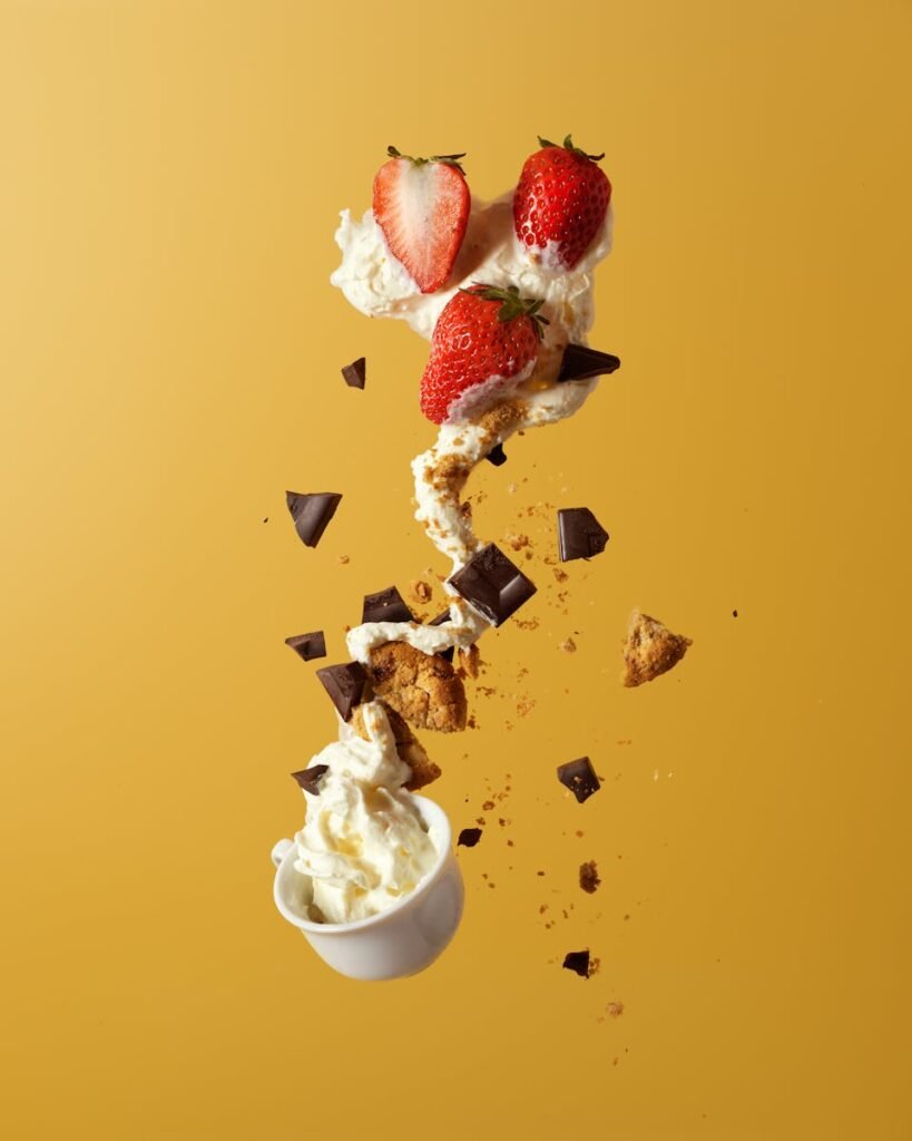 Delicious strawberries and cream with chocolate and cookie pieces, captured in mid-air against a vibrant yellow background.